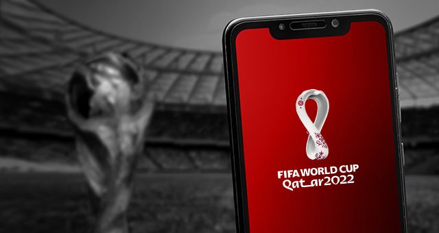 a smartphone with the FIFA World Cup Qatar 2022 logo on the screen and a football stadium and the World Cup trophy in the background